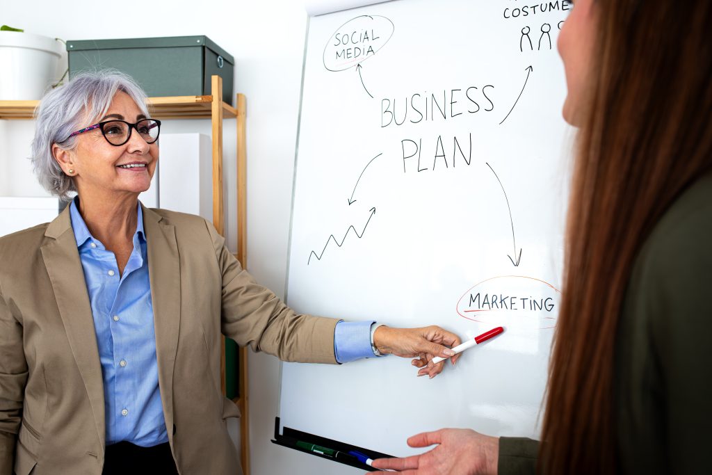A woman points to a whiteboard displaying a business plan titled "Why Website Rebuilds Fail without Business Planning."